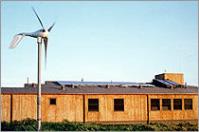 Photo of a wood building with a small wind turbine in front and photovoltaic panels on the roof.