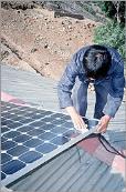 A photo of a technician inspecting a photovoltaic array on the roof of a house. The photovoltaic array contains many square-shaped, black solar cells contained beneath translucent glass.
